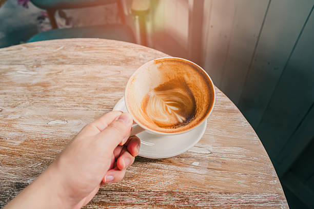 Hand Holding Coffee Cup with Swirling Foam on Rustic Wooden Table in Bright Cafe Setting