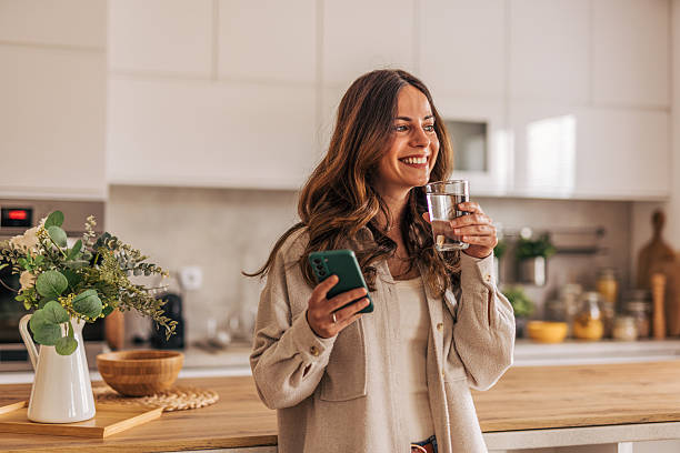 istock Smiling woman drinking water and using smartphone in kitchen 2218322646