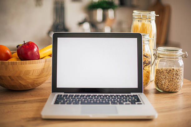 Laptop with blank screen is placed on kitchen table with fruits and cereals, suggesting online recipe or healthy lifestyle