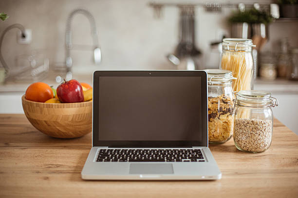 Laptop with blank screen placed on kitchen table next to a bowl of fresh fruit and jars of cereals
