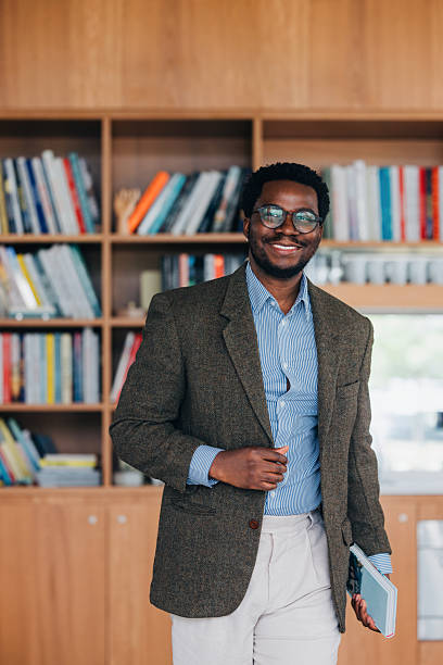 A well-dressed man stands confidently in a contemporary library setting, holding a book. The scene conveys professionalism, education, and positivity, with bookshelves filled with colorful books enhancing the background.