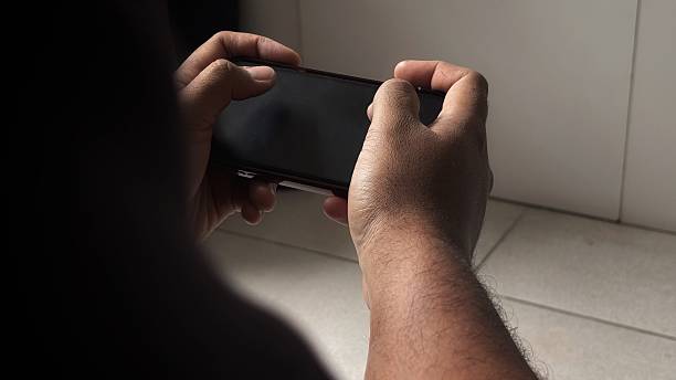 This image captures a person engrossed in a mobile game, seen from over their shoulder. The individual is holding a smartphone horizontally with both hands, thumbs positioned on the screen, indicating active gameplay. The screen itself is dark, suggesting either a dark game interface or the phone is off. The setting appears to be indoors, with a subtle light source from the right illuminating parts of their hands and a white wall in the background. The focus is clearly on the interaction between the user and their device, highlighting the common act of mobile gaming.