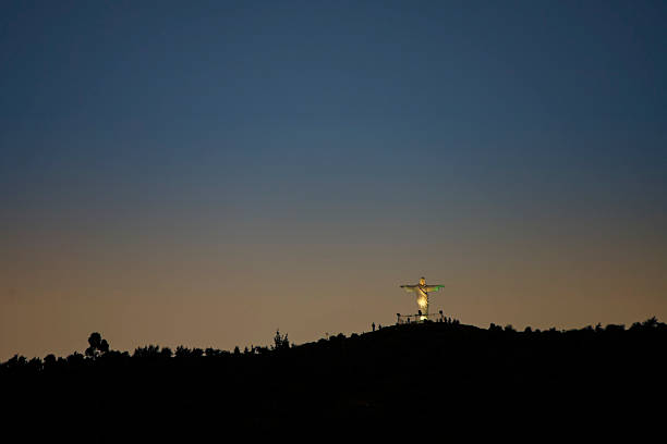 Statue of Christ at sunset at the top of a hill overlooking the city of Cusco, Peru down below.  Photo taken shortly after sunset with orange glow fading to dark blue sky above.