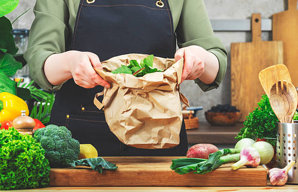 istock Chef holds paper package with vegetables peels for processing compost. Wooden table, kitchenware, vegetables and herbs. Healthy eating, zero waste, blogging, recycling 2218338508