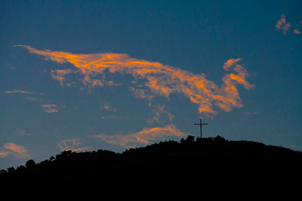 Cross on top a hill and in silhouette against the sky, above Cusco, Peru at sunset, wispy orange clouds in the dark blue sky.