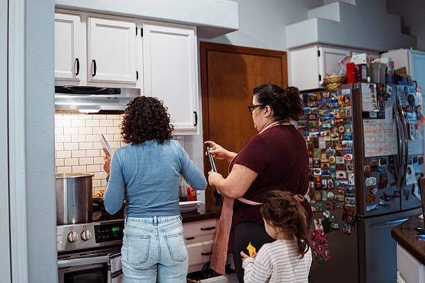 A Mexican woman who is hosting friends for dinner and prepared beans and tamales, chats with an Eurasian friend as the woman serves herself and her daughter food from the stove.