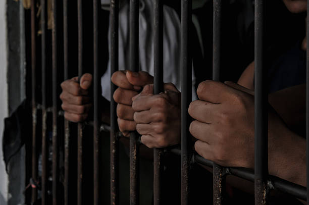 A close-up of hands gripping prison bars creates a powerful image of confinement. The dramatic lighting enhances the emotions of despair and isolation.