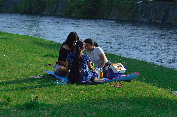 Seated by a gently flowing river, three teenage girls laugh and enjoy a calm conversation during their morning picnic.