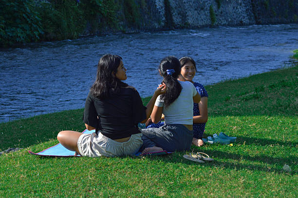 The girls sit on a mat next to a calm river, with one of them turning back as if responding to a sound or enjoying the view behind