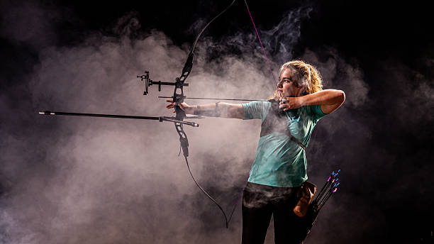 A female archer powerfully draws her bow in a dramatic, smoke-filled studio setting, embodying determination, focus, and athletic precision.