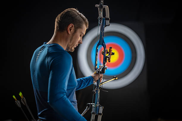 A male archer in a blue shirt lowers his compound bow and looks down thoughtfully after shooting, standing before a bullseye target in a low-lit studio setting.
