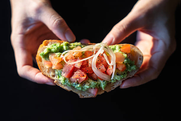 Close-up of hands holding a slice of fresh avocado toast with diced tomatoes and onions on a rustic bread slice, ready to eat.