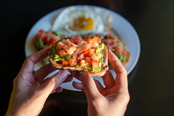 Overhead view a person hands holding a slice of fresh avocado toast with diced tomatoes and onions on a rustic bread slice, ready to eat.