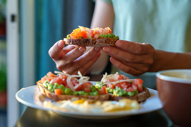 Close-up of hands holding a slice of fresh avocado toast with diced tomatoes and onions on a rustic bread slice, ready to eat.
