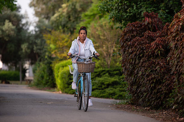 A woman rides a classic blue bicycle down a peaceful, leafy road bordered by dense green and red foliage. Dressed in pastel tones with a light jacket, jeans, a white t-shirt, and a pink knitted scarf, she embodies comfort and style. The bicycle includes both a front wicker basket and a rear metal basket, enhancing the image’s charm and practicality. Captured in soft daylight, this scene evokes a tranquil, eco-conscious lifestyle ideal for themes of urban serenity, personal wellness, and slow living.