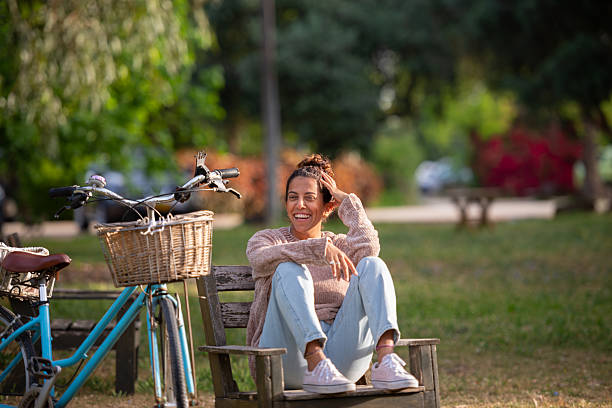 A cheerful woman sits casually on a wooden park bench, smiling and enjoying the sunshine. She is dressed in light denim jeans, a soft knitted sweater, and white sneakers. Her vintage blue bicycle, featuring a front wicker basket, is parked nearby, enhancing the relaxed and carefree atmosphere. The lush greenery and vibrant flowers in the background evoke a warm, peaceful spring setting — ideal for themes such as slow living, wellness, outdoor leisure, and urban nature.