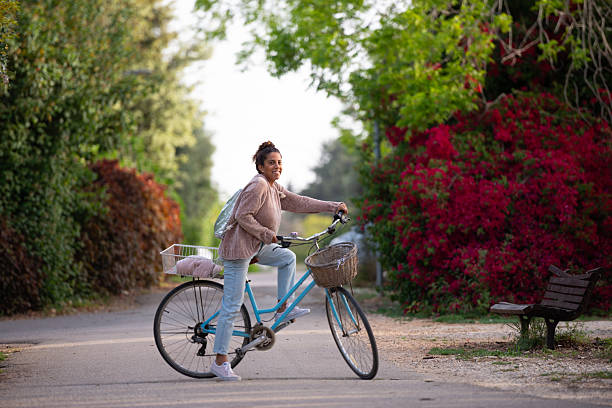 A woman prepares to ride her vintage blue bicycle along a beautiful tree-lined path, surrounded by lush greenery and vivid red flowering bushes. She wears light blue jeans, white sneakers, and a cozy pinkish-brown sweater, with a metallic backpack and a pink scarf in the rear basket of the bike. The soft lighting and bright foliage create a warm, peaceful, and cheerful atmosphere, perfect for themes of springtime, nature, wellness, and sustainable living.