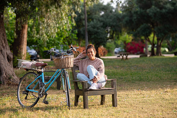A woman sits comfortably on a wooden bench in a sunny park, smiling and enjoying the moment. She wears light-wash jeans, white sneakers, and a cozy pink-beige sweater. Her vintage blue bicycle with a wicker front basket and metal rear basket is parked beside her, creating a warm, serene scene. Surrounded by green trees and soft natural light, the image captures the essence of outdoor leisure, wellness, and joyful solitude.