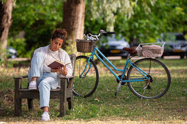 A young woman sits cross-legged on a rustic wooden bench in a sunny park, deeply focused on reading a book. She wears light blue jeans, a white t-shirt, and an open plaid shirt. Next to her stands a vintage blue bicycle with a wicker basket in the front and a blanket in the rear basket. The background of green trees and parked cars creates a relaxed, natural atmosphere, conveying a sense of calm, reflection, and everyday life outdoors.