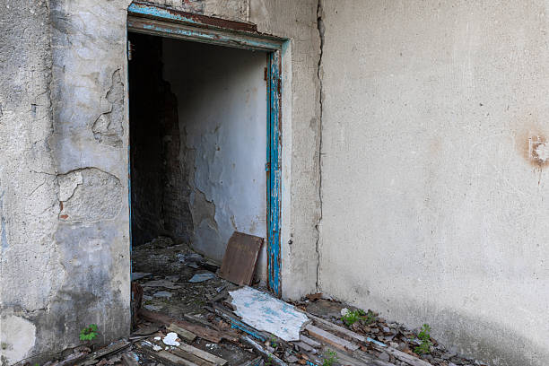 Weathered doorway in a decaying concrete structure with peeling paint, exposed brick, and debris-covered floor suggesting neglect and abandonment.