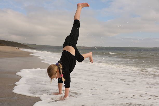 Young blonde haired girl performing a handstand in breaking waves on a summer UK beach holiday. She is wearing black clothing