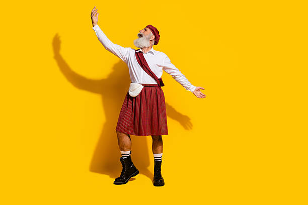 A senior man dressed in traditional Scottish clothing poses dynamically, gesturing expressively on a vibrant yellow background, evoking cultural celebration and lively charisma.