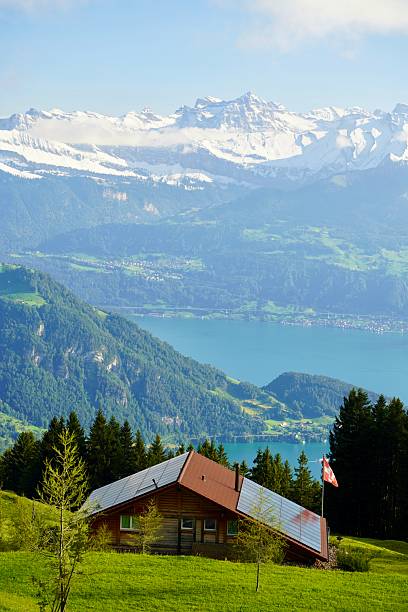 Scenic view of a traditional wooden cabin with solar panels and a Swiss flag, set on a lush green hillside. The cabin overlooks a turquoise alpine lake, with majestic snow-covered mountains rising in the background under a clear blue sky. This tranquil setting captures the essence of sustainable living, adventure travel, and the natural beauty of the Swiss Alps or a similar mountainous region.