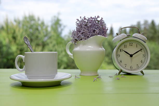 A white cup of coffee stands next to a retro alarm clock and a white jug of lilac flowers on a green wooden desk against a summer woodland background.