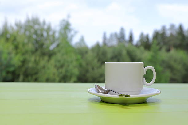 A white cup of coffee stands on a green wooden desk against a summer woodland background.