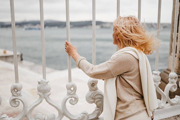Portrait of beautiful mature blond woman standing and looking at sea through fence of Dolmabahce Palace in Istanbul on windy overcast day