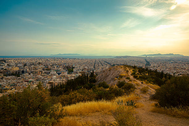 A panoramic view of Athens, Greece, captured from the scenic Philopappos Hill during the golden hour. The image reveals a contrast between urban density and natural landscape, with the Aegean Sea and distant mountains visible on the horizon. The walking path in the foreground winds through Mediterranean vegetation, leading the eye toward the sprawling city below. Ideal for use in travel editorials, landscape design projects, tourism campaigns, or visuals exploring the intersection between nature and urban development.