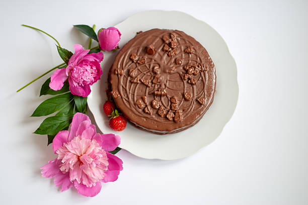 Top view of a round chocolate cake with decorative frosting and chocolate chunks, served with fresh strawberries and arranged beside blooming pink peony flowers on a white plate. Set against a clean white background, this composition conveys elegance and celebration. Ideal for themes like homemade desserts, festive occasions, summer sweets, or romantic settings.