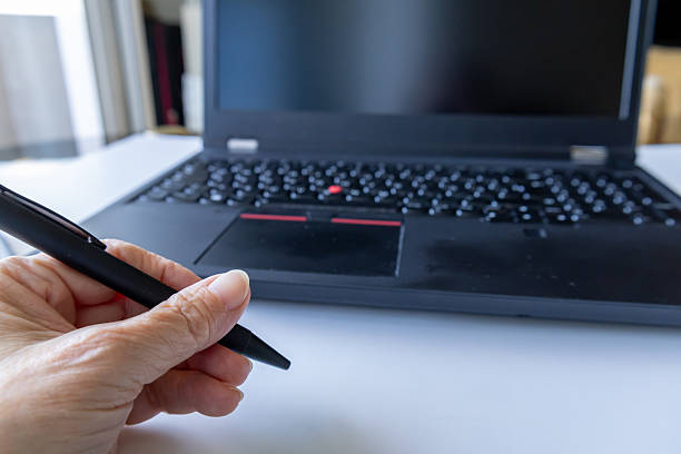 A black laptop on a desk with a hand holding a black pen next to it. The image suggests work, creativity, or planning in a modern environment, combining digital and traditional tools for productivity.