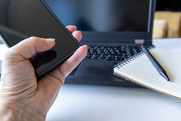 An elderly hand holds a modern smartphone in front of a black laptop and an open notebook with a pen. The image captures a multitasking work environment, blending digital and analog tools. The soft lighting and sharp focus emphasize the contrast between the textured skin and sleek devices.