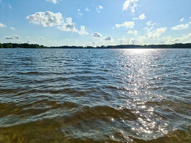 Sun reflecting on lake water surface with wind turbines and forest in background, on a beautiful summer day with blue sky and white clouds