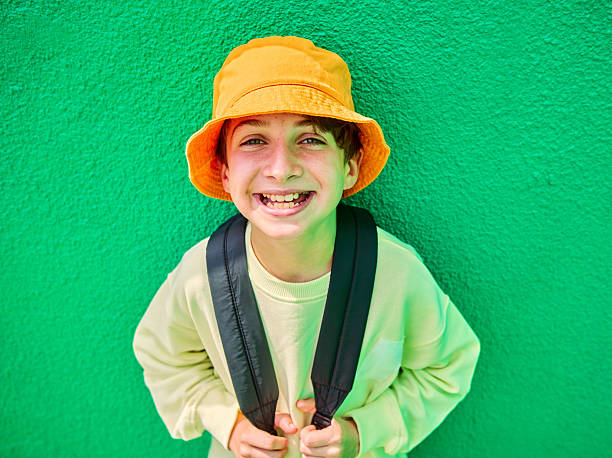 Portrait of a cheerful student wearing an orange bucket hat and backpack, smiling brightly against a vibrant green wall