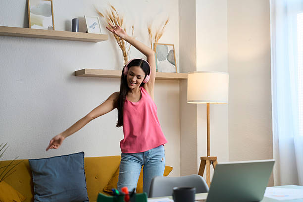 Happy young woman dancing while working from home and listening to music with headphones