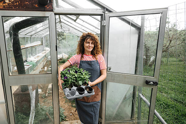 istock Young beautiful woman posing for camera while holding potted plants and standing in front of green house 2218437890