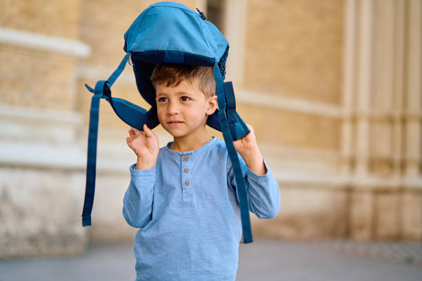 Preschool age child joyfully wearing a backpack on his head while playing in front of a school building, enjoying a sunny day filled with laughter and carefree exploration