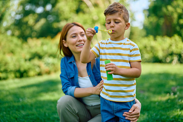 Happy mother and son enjoying playful moments while blowing soap bubbles together in a lush green park on a bright, sunny day, surrounded by nature and laughter