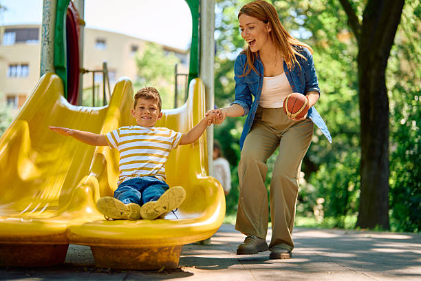 Happy mother holding her son's hand while he joyfully slides down the bright yellow slide at the playground, soaking up the sunshine on a delightful summer day outdoors