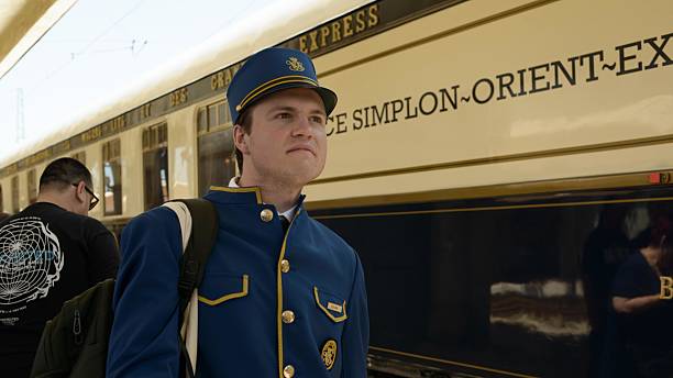 June 03,2025  - Ruse, Bulgaria: A uniformed Venice Simplon Orient Express steward in blue and gold stands by the cream and navy historic carriage, symbolizing premium hospitality and Art Deco luxury on European rail journeys.