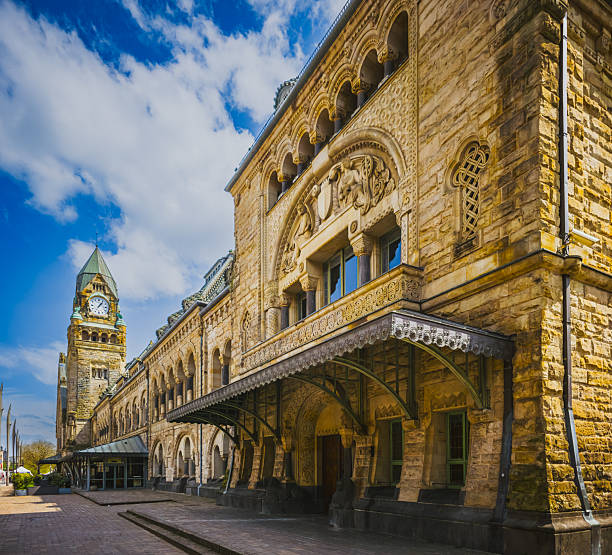 Exterior view of the Gare de Metz-Ville train station in Metz, France. The photo highlights the historic Romanesque revival architectural details, including the ornate stone facade, clock tower, and arched windows, captured on a sunny day with blue skies.