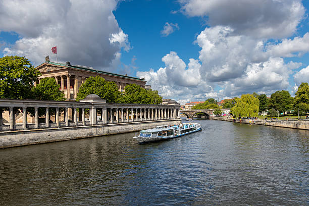 Berlin, Germany, May 26, 2025: A modern sightseeing boat sails along the Spree River, passing Museum Island landmarks and lush greenery under dramatic clouds.