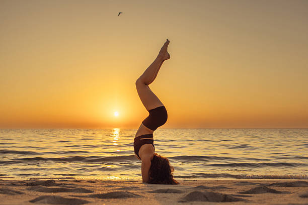 A person practices a yoga pose on a sandy beach as the sun rises over tranquil waters. The calm and serene atmosphere complements the focus and balance required for the exercise, creating a peaceful and inspiring scene.