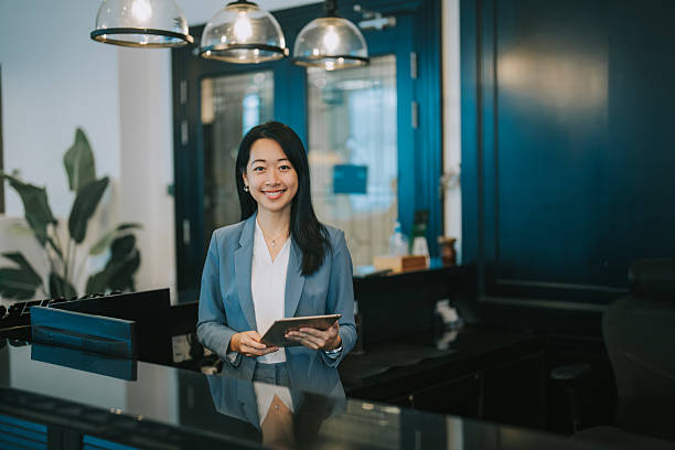 A smiling young Asian woman in professional attire stands at a modern office reception counter, holding a digital tablet. The environment is sleek and contemporary, conveying professionalism, confidence, and digital workplace readiness. Ideal for concepts like leadership, modern office, receptionist, or corporate services.