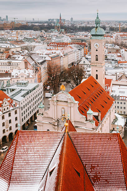 High-angle winter view of Munich, taken from the top of St. Peter’s Church tower. Snow blankets the rooftops of historic buildings, creating a picturesque winter cityscape. The scene overlooks Tal street, St. Peter Church’s rooftop (12th century), and the facade and tower of Heilig Geist Church (14th century), capturing the timeless charm of Munich’s old town.