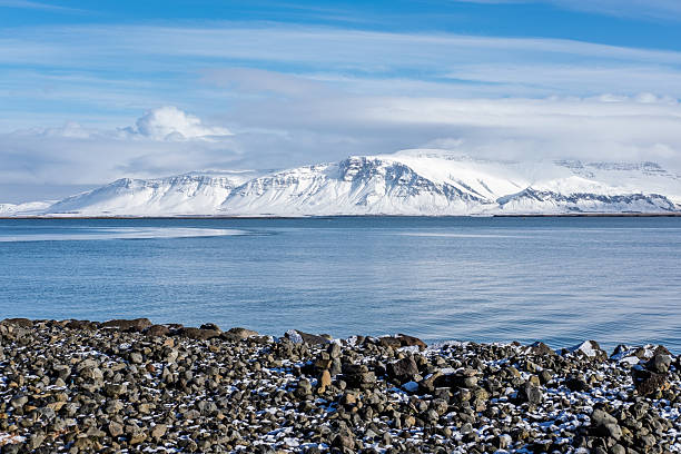 istock This image depicts a serene winter landscape. Snow-covered mountains form the backdrop to a calm body of water and a rocky shore dusted with snow. 2218486531