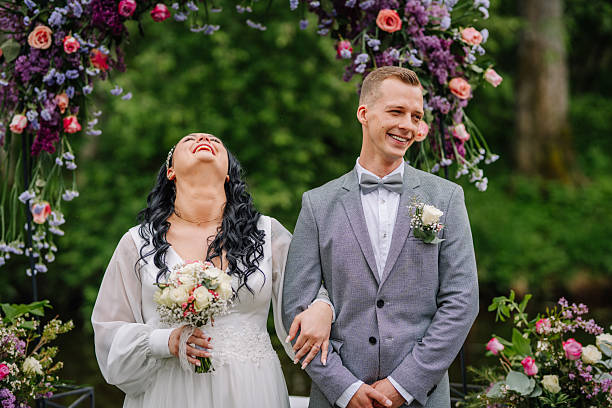 Bride and groom share a joyful moment under a floral arch during their wedding ceremony, laughing and smiling in a lush garden setting.