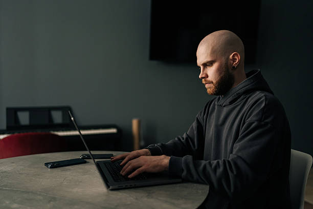 istock Side view of focused male programmer wearing dark hoodie typing code on laptop computer, sitting at table near window in contemporary urban apartment. 2218490201